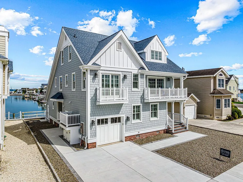 A multi-story gray shingled coastal house with white trim, a garage, and balconies, situated near a canal under blue sky.