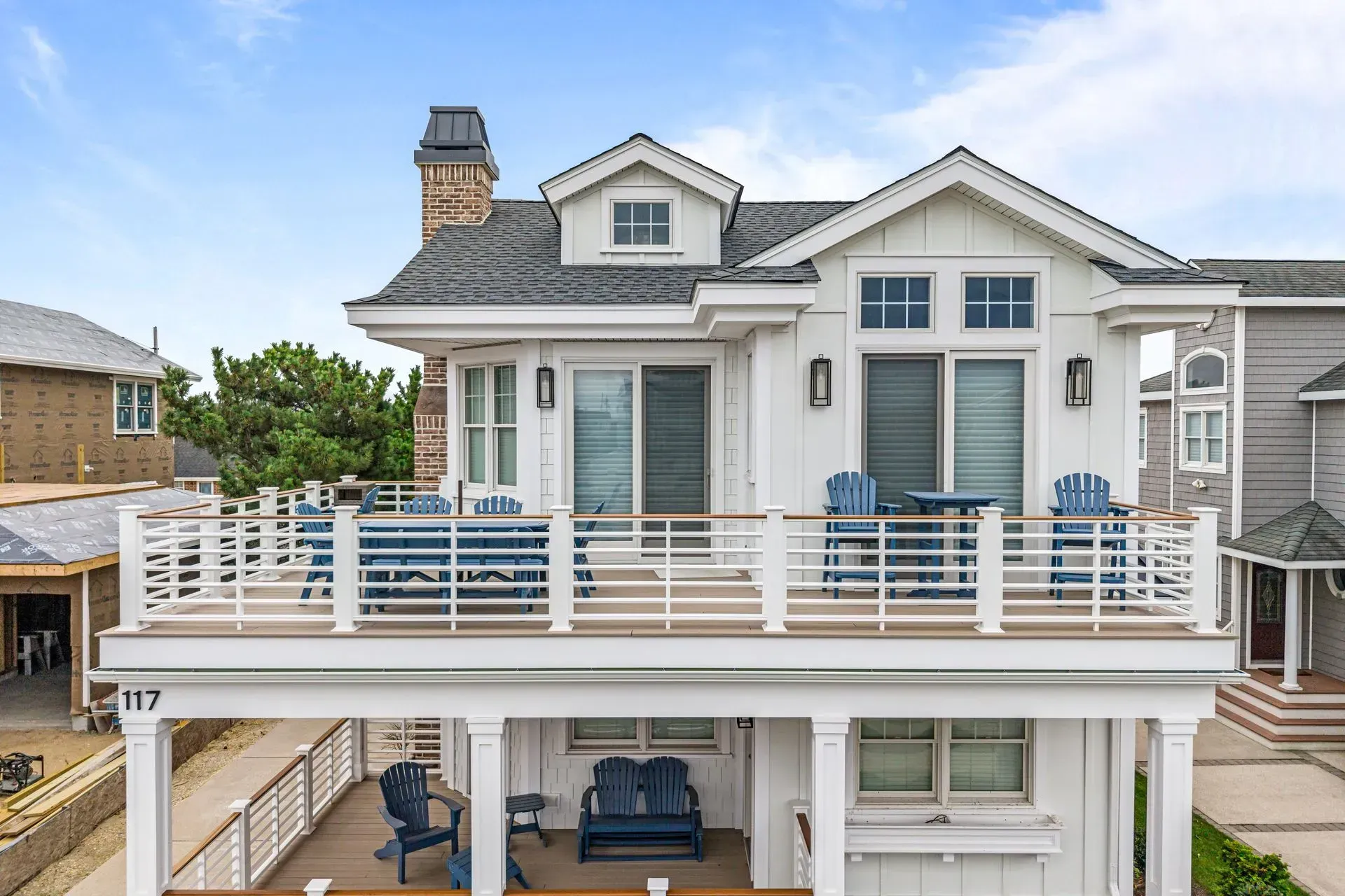 A two-story white beach house with a prominent second-floor deck, outdoor seating, a brick chimney, and a blue sky.