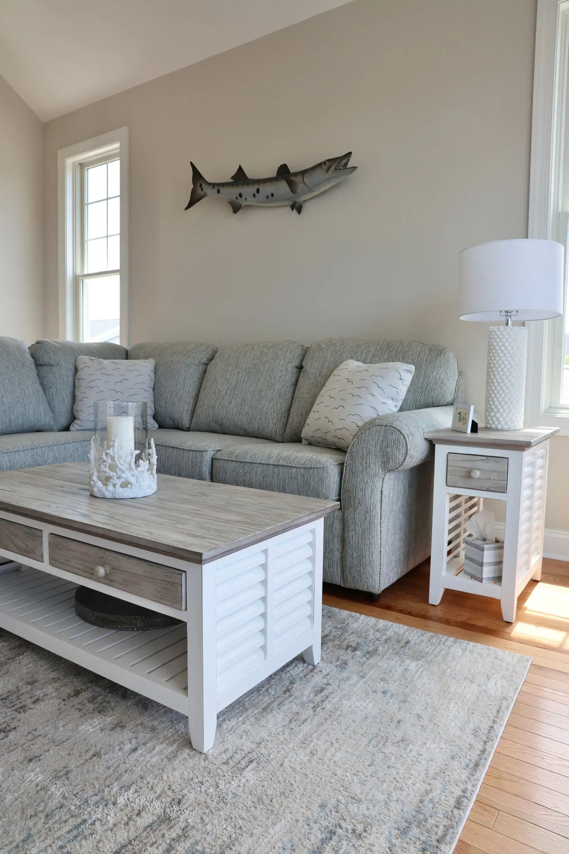 A living room with a patterned grey sectional sofa, white coffee table, matching side table, and a fish wall ornament.