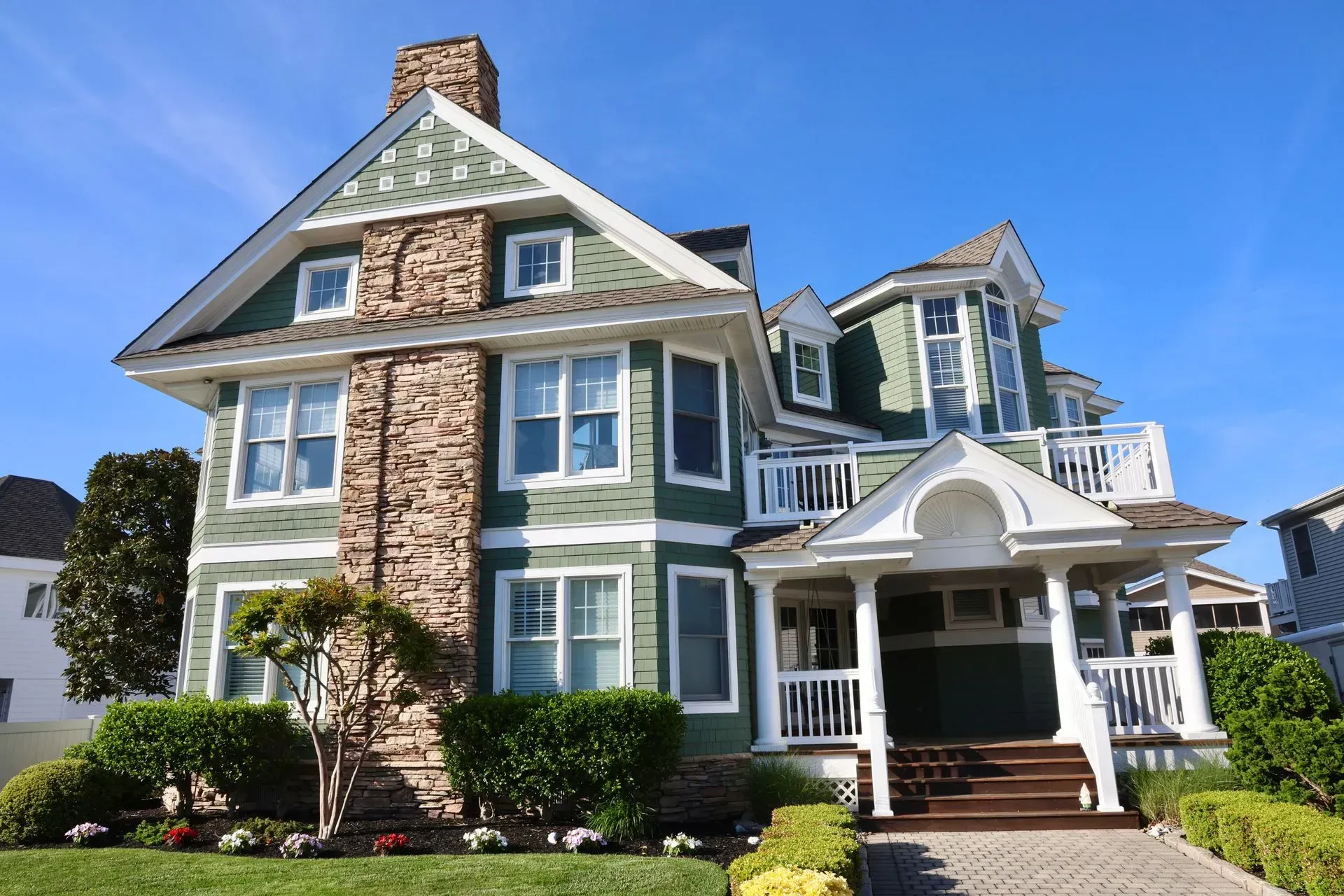 A green Victorian-style house with a stone chimney, white porch, and lush landscaping against a clear blue sky.