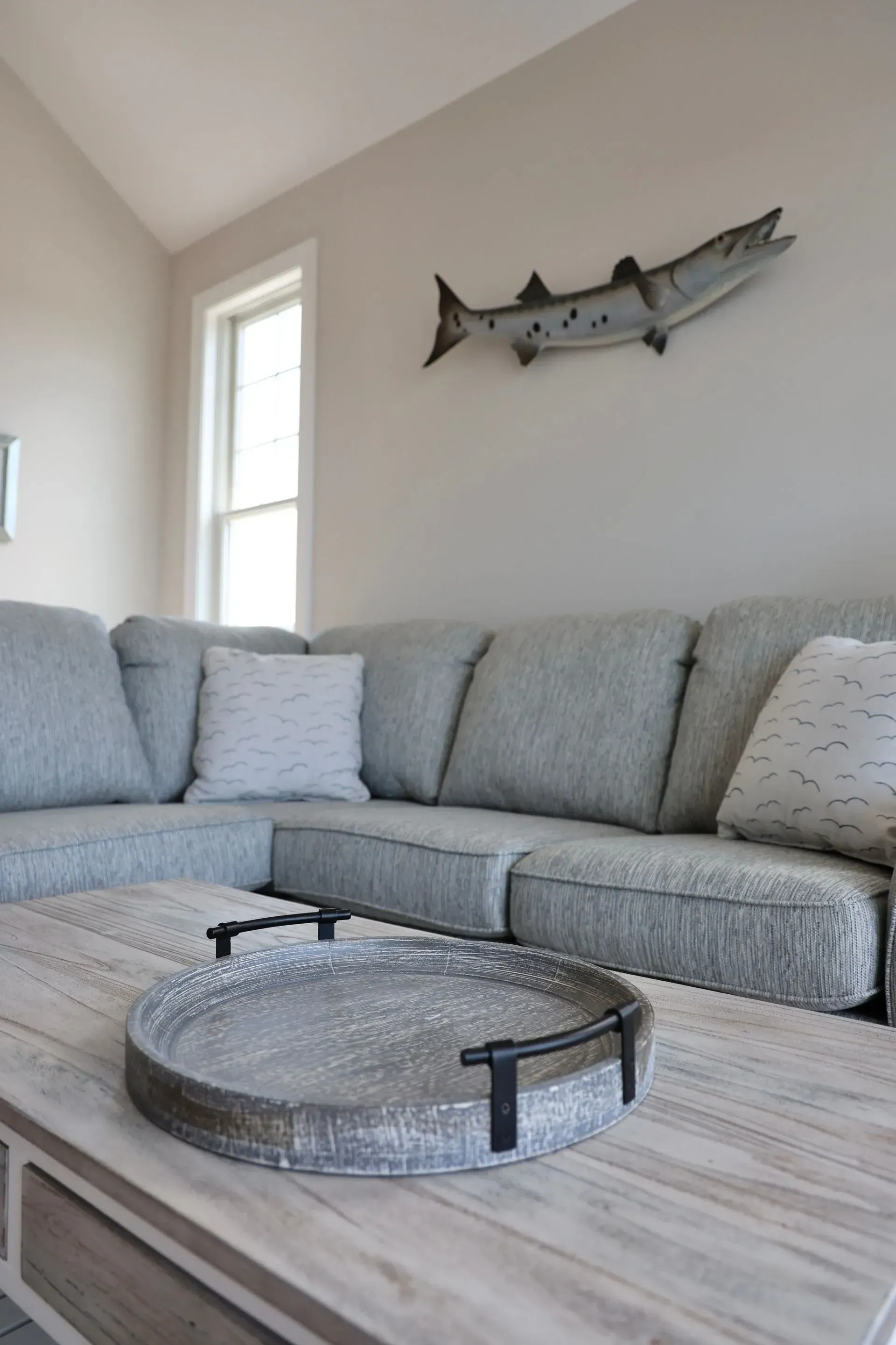 A gray sectional sofa and a weathered wood coffee table with a round, textured tray, beneath a fish wall sculpture.