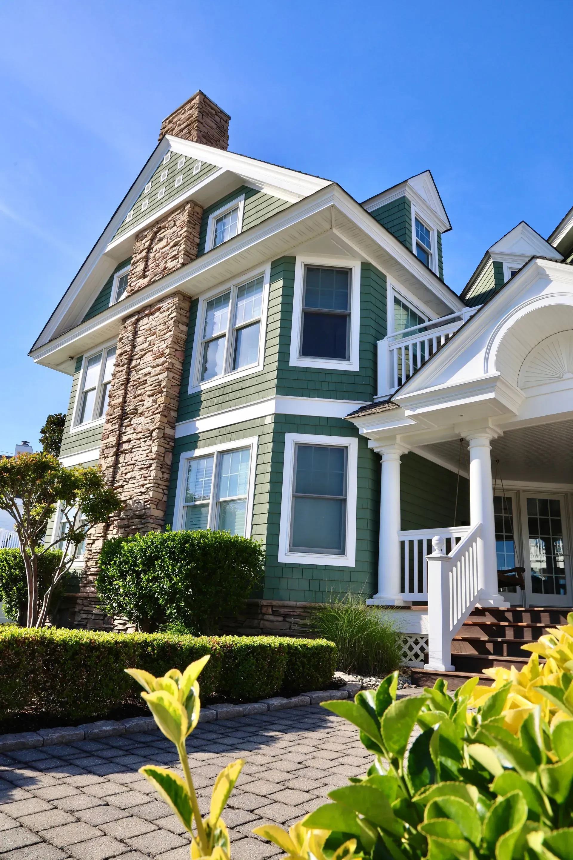 A three-story green house with stone chimney, white trim, and a porch, framed by a bush in the foreground under blue sky.