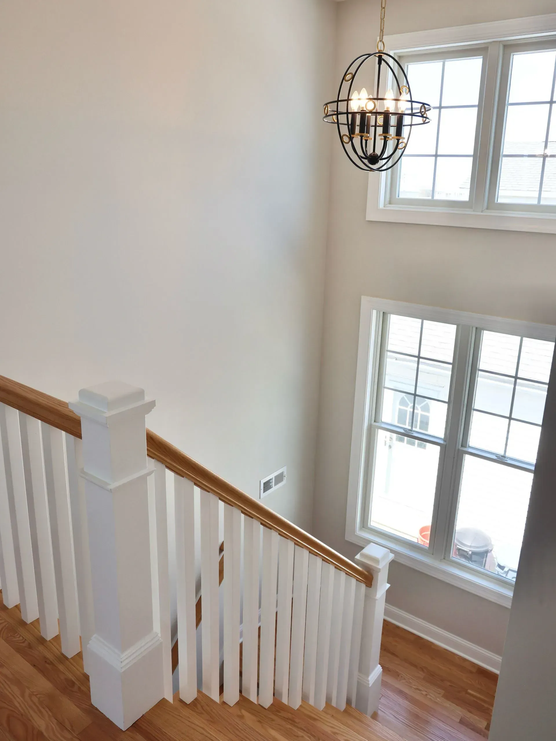 A staircase with white balusters and wood handrails, illuminated by a hanging orb chandelier and two-story tall windows.