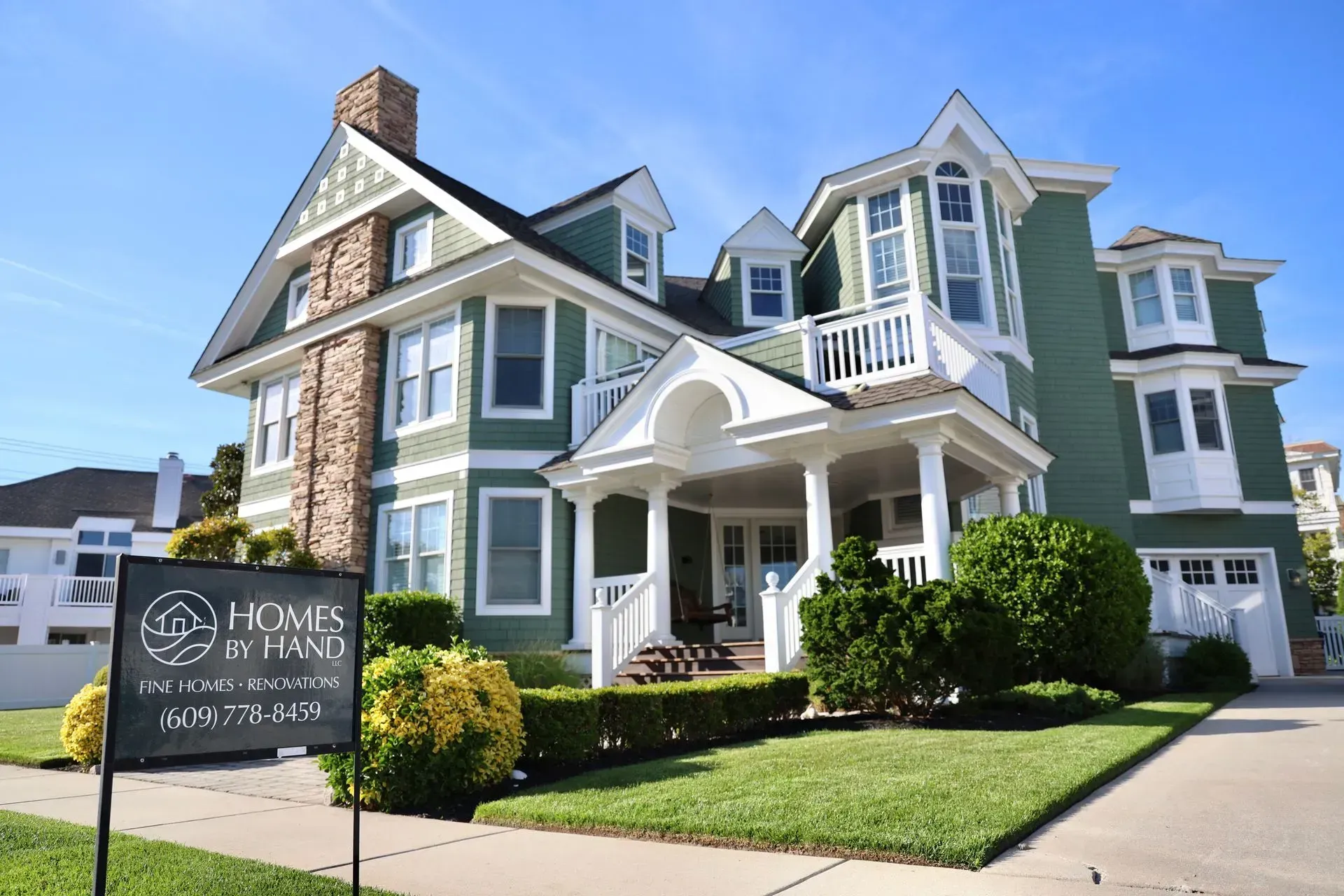A large green multi-story house with white trim, stone accents, and a front yard, viewed from the street with a real estate sign.