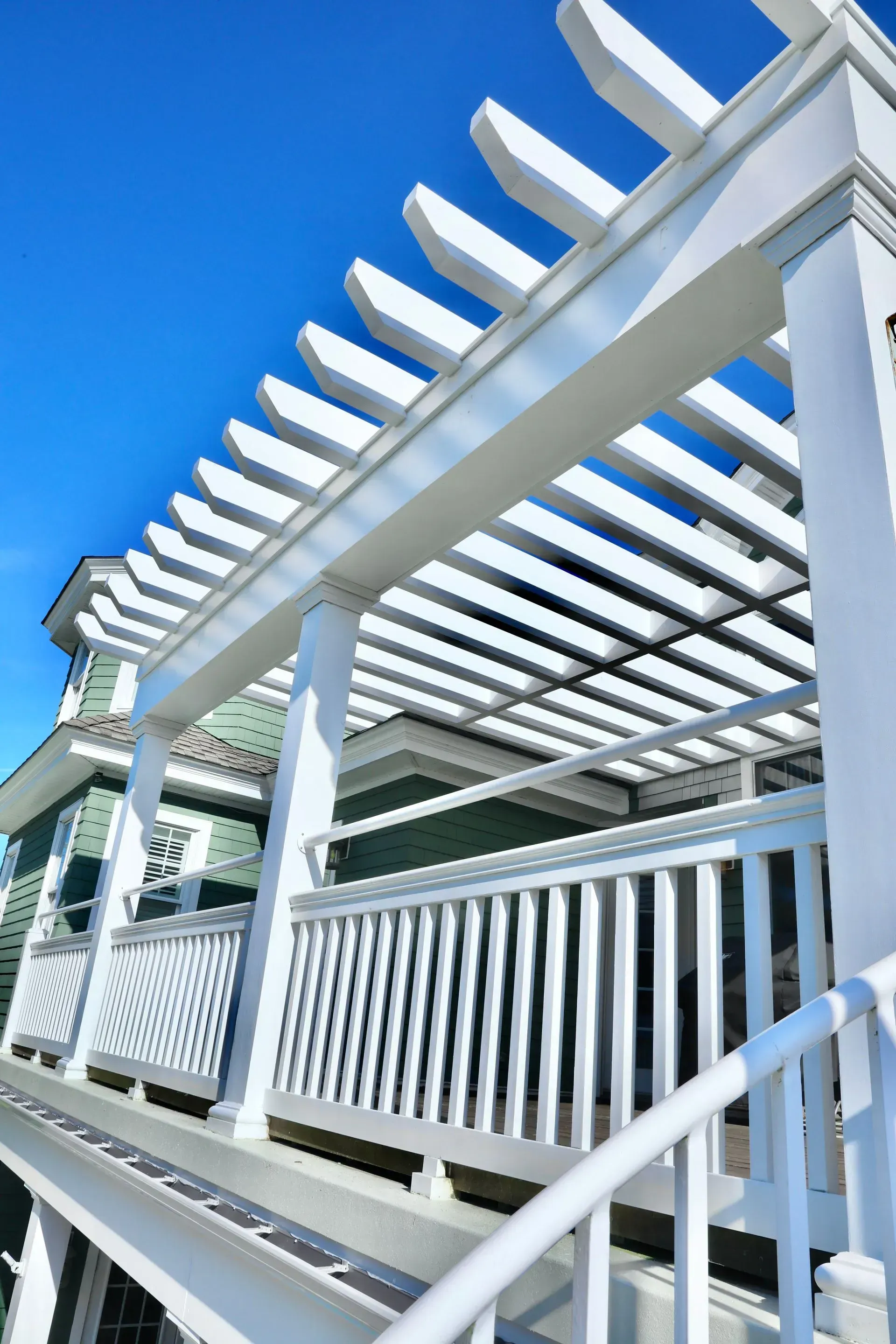 A white wooden pergola attached to a green house with a balcony railing against a bright blue sky.