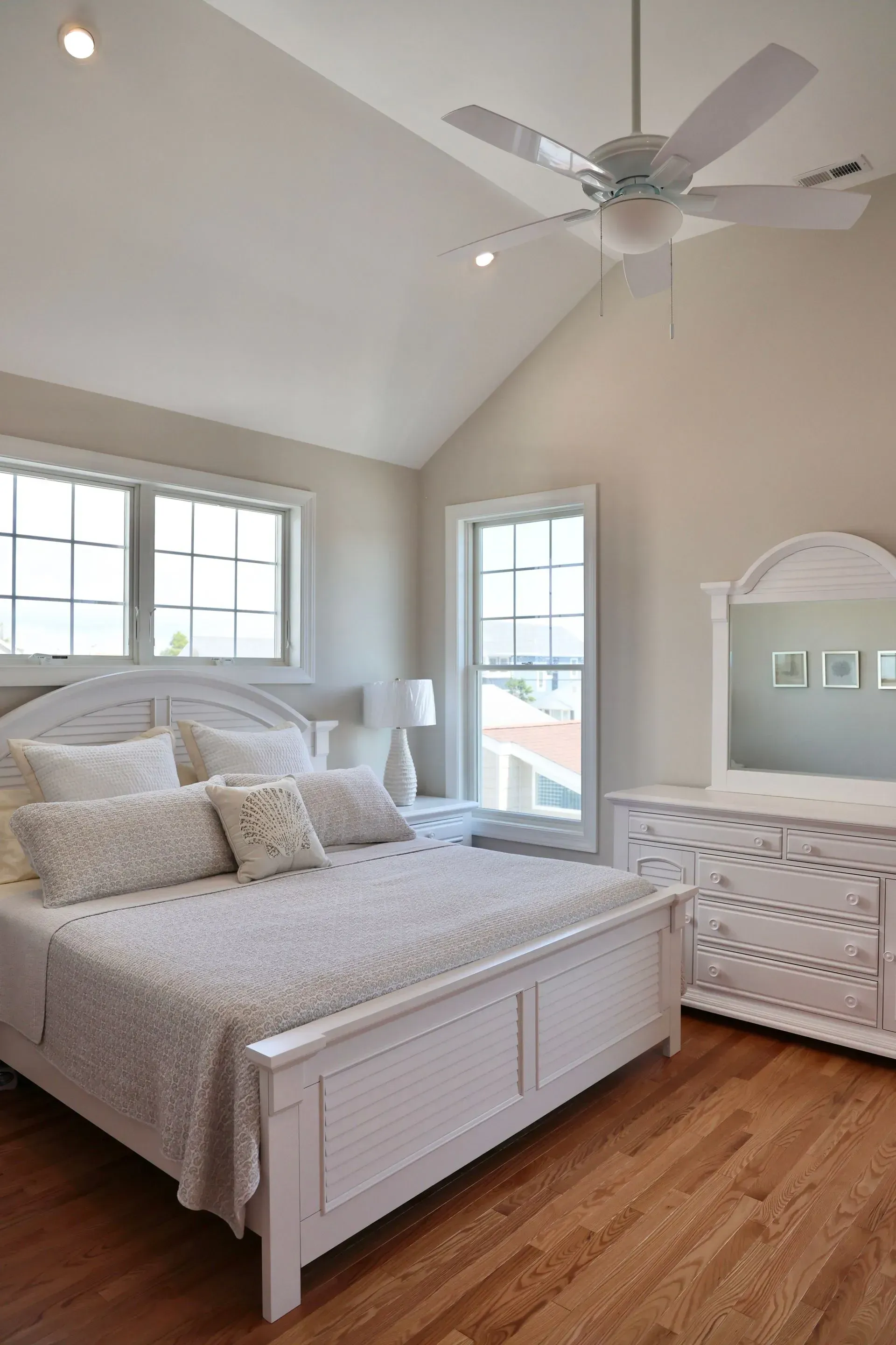 A bedroom with a white bed, white dresser with mirror, hardwood floors, large windows, and a ceiling fan.