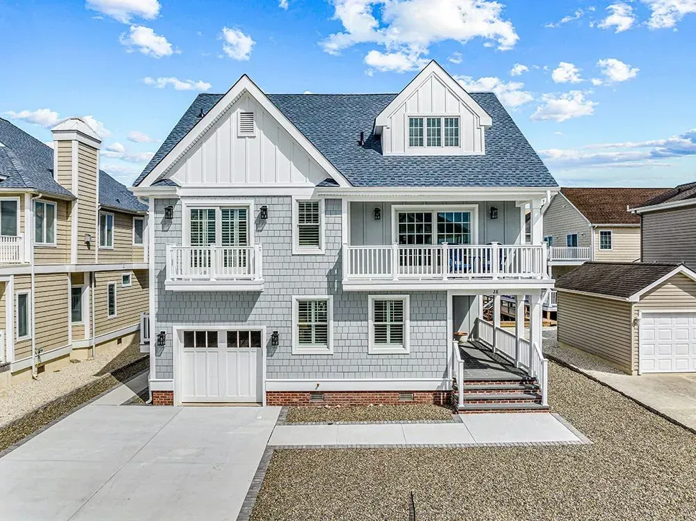 A light gray, two-story beach house with a white garage, front balconies, a blue shingled roof, and a gravel driveway.