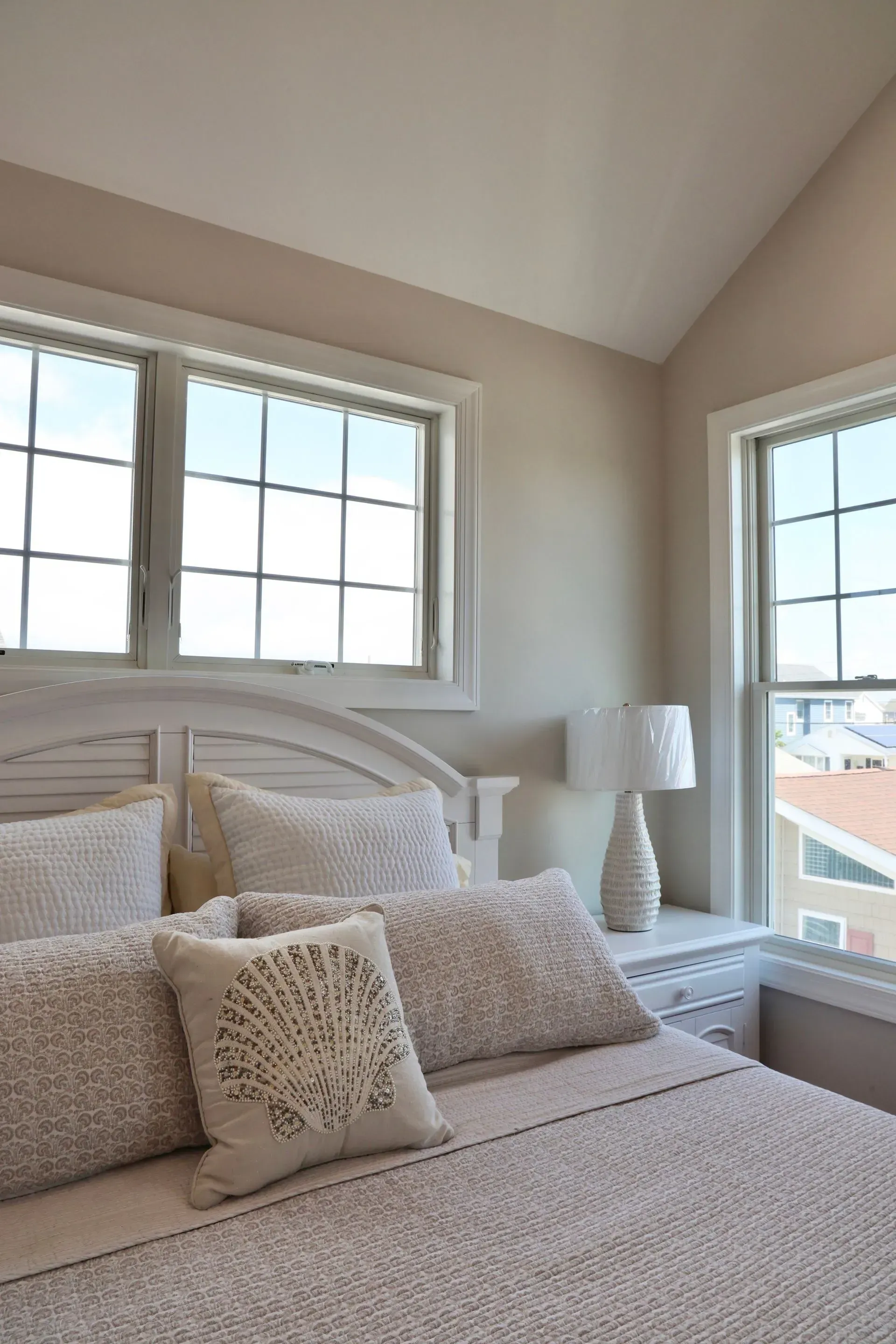 A white bedroom with a bed, a decorative shell pillow, a bedside lamp, and windows with a view of nearby houses.