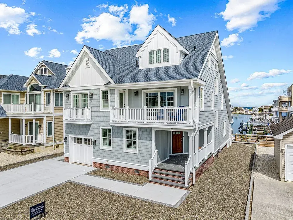 A light-gray coastal house with a blue roof, white trim, and wrap-around balconies, situated near a marina under blue sky.
