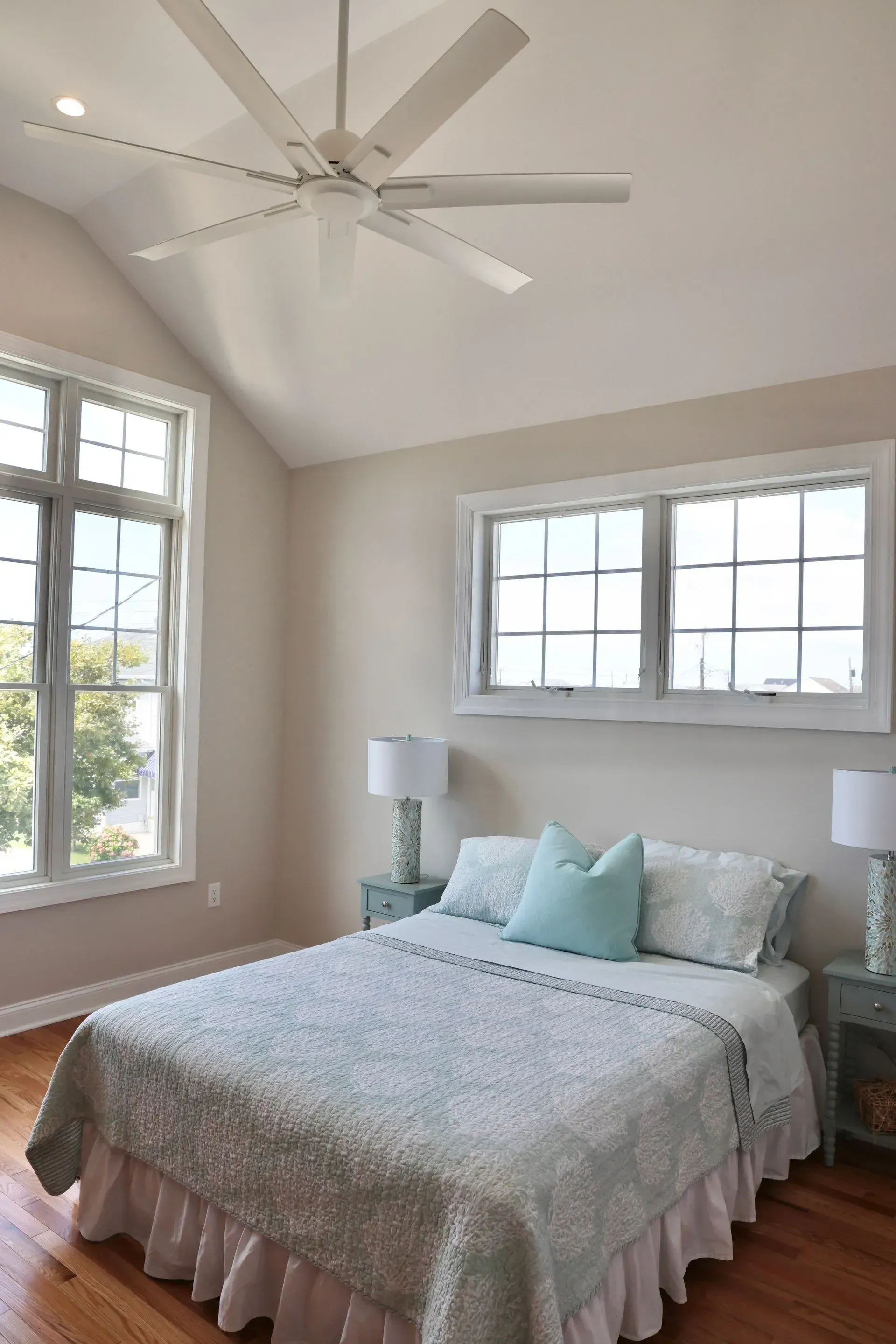 A bedroom with a bed, two nightstands, a vaulted ceiling with a fan, and windows on a neutral-colored wall.