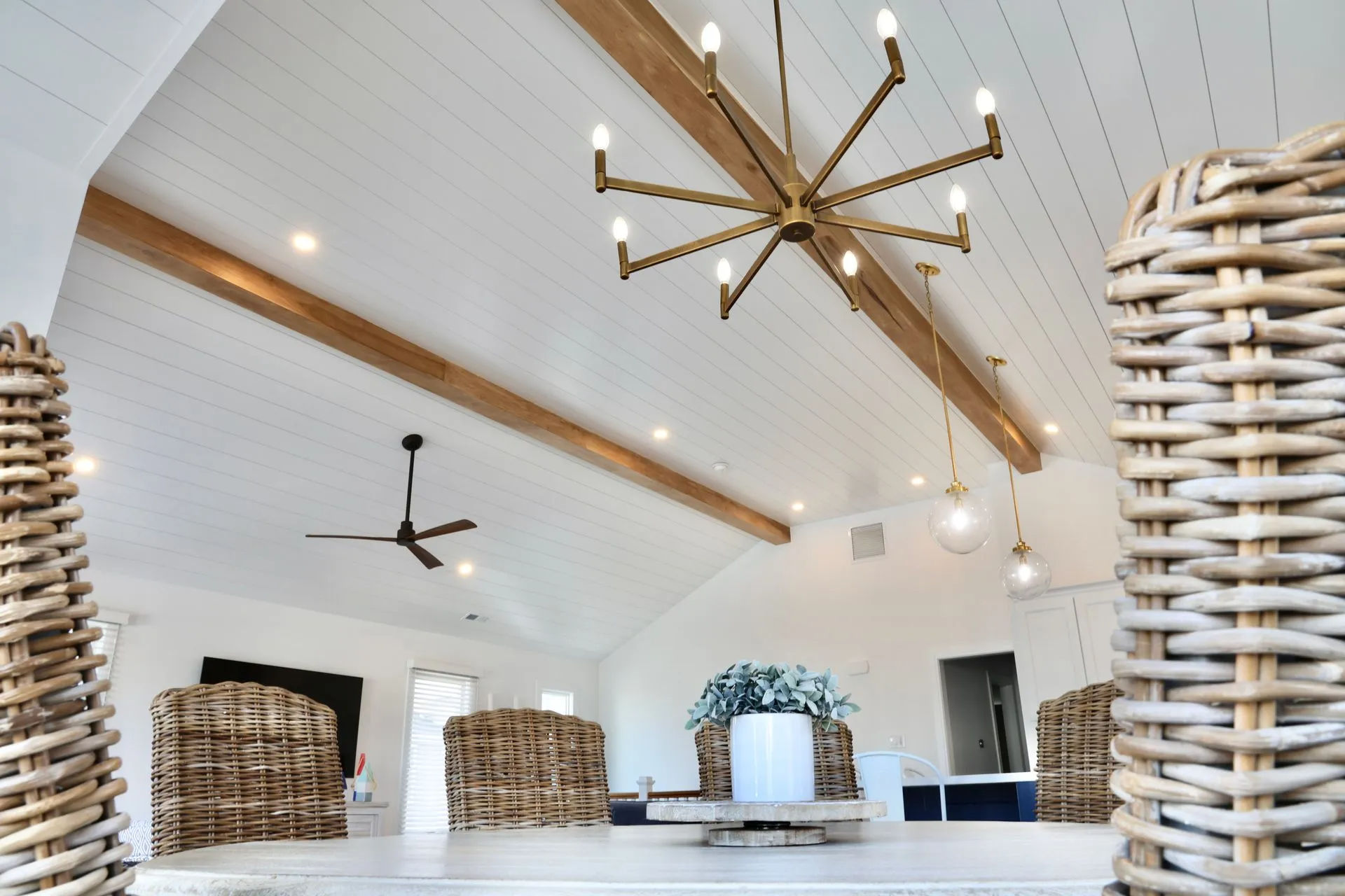 A dining area with woven chairs, a white table, a large gold chandelier, and wooden beams on a white shiplap ceiling.