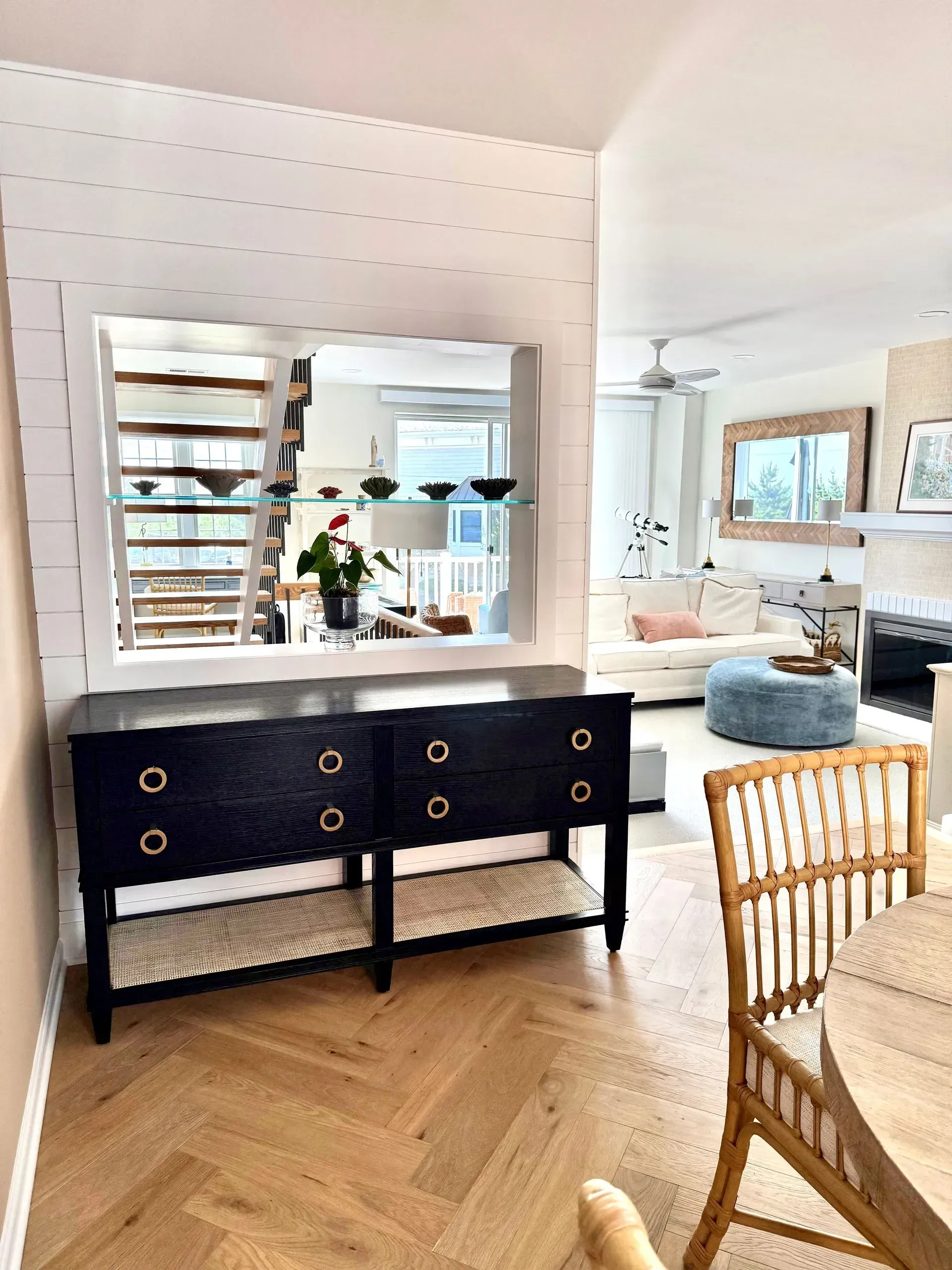 A black cabinet with brass ring pulls sits in front of a shiplap wall featuring a window looking into an adjacent room.