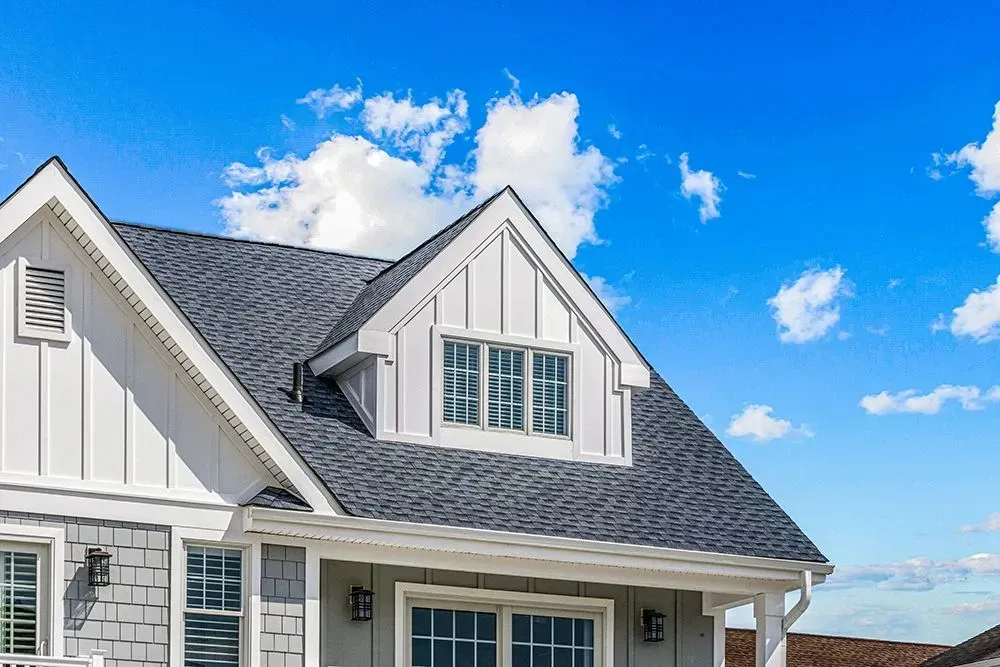 A white house with gray shingle roofing and a dormer window under a bright blue sky with white clouds.