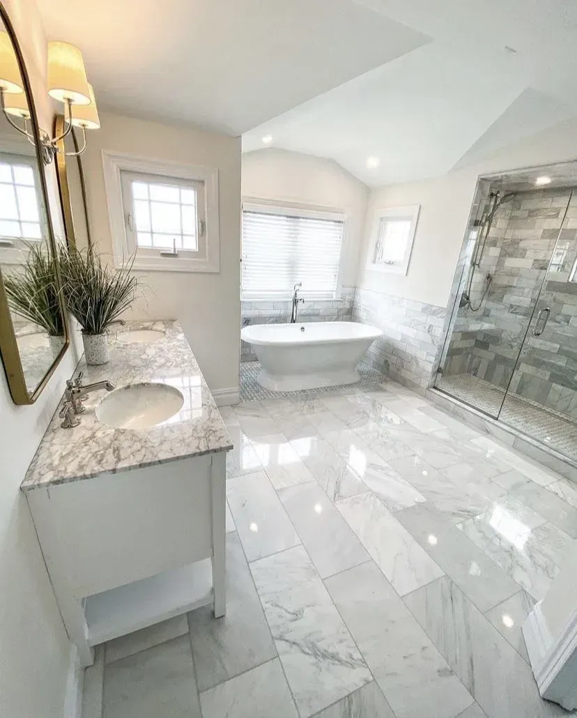 A bright, modern bathroom featuring white marble tile, a pedestal sink, a freestanding tub, and a glass-enclosed shower.