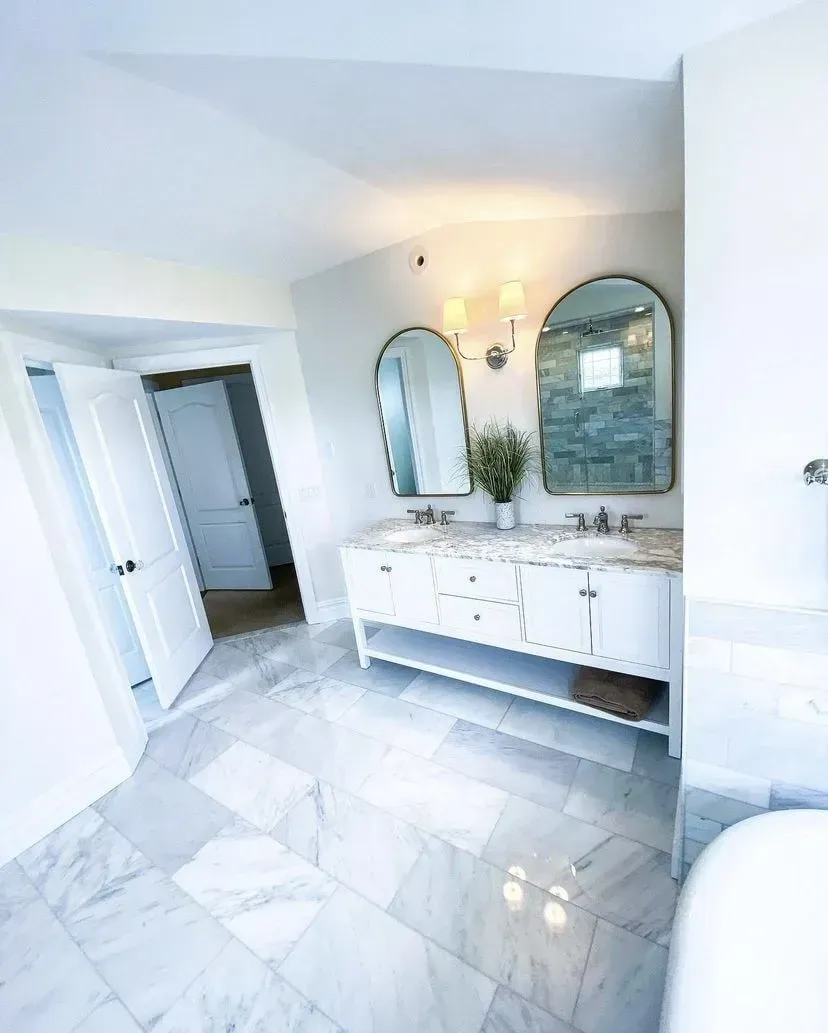 A bright, modern bathroom with a double white vanity, arched mirrors, and white marble tiled floors.