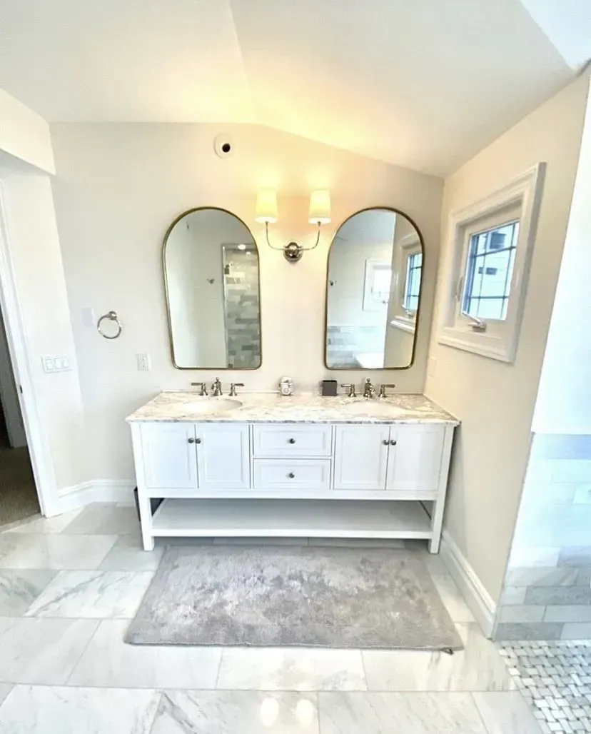 A double-sink bathroom vanity with white cabinets, marble countertops, two arched mirrors, and a gray rug on tiled floor.