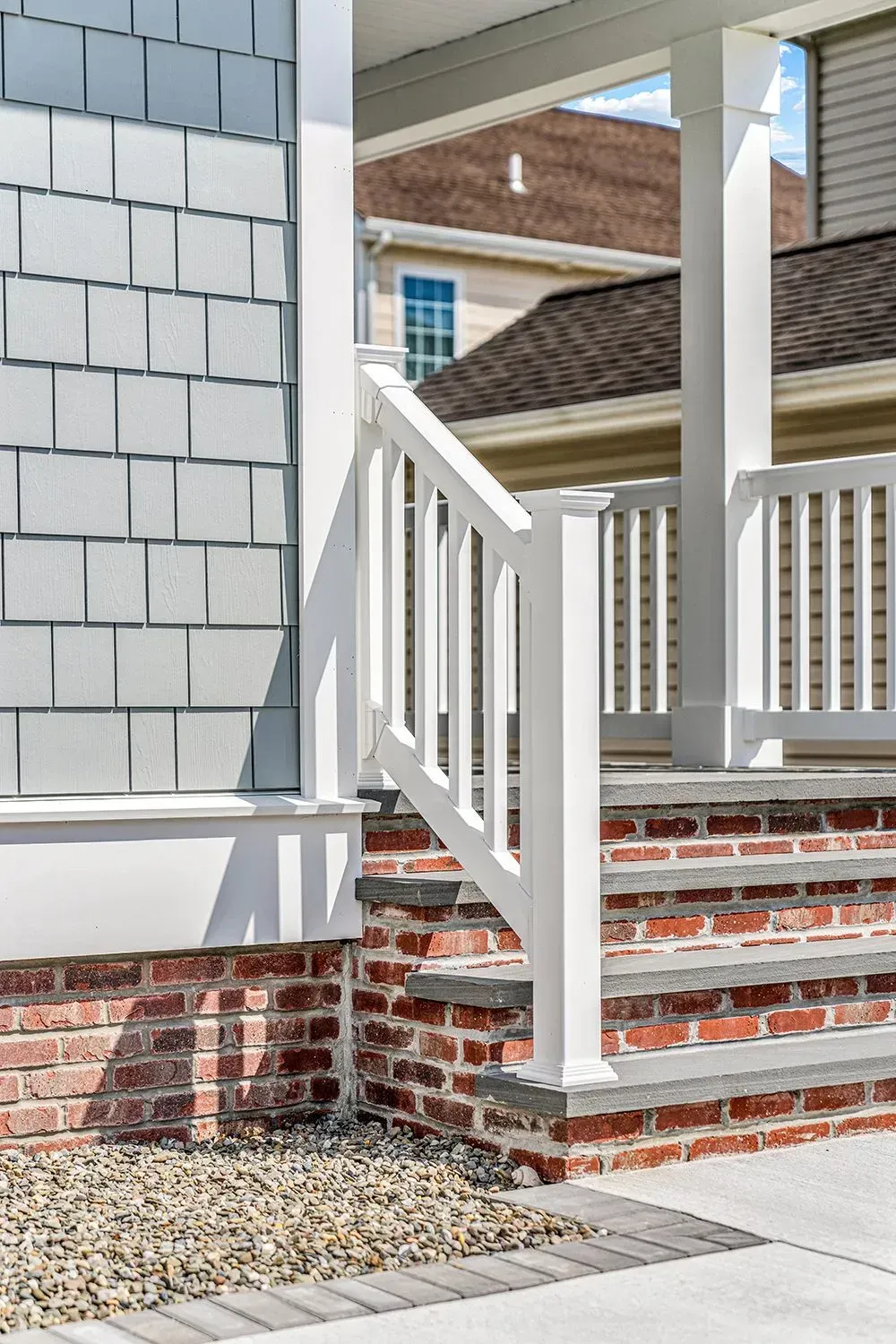 White porch steps with a railing leading to a house exterior featuring gray shingles and a brick foundation.
