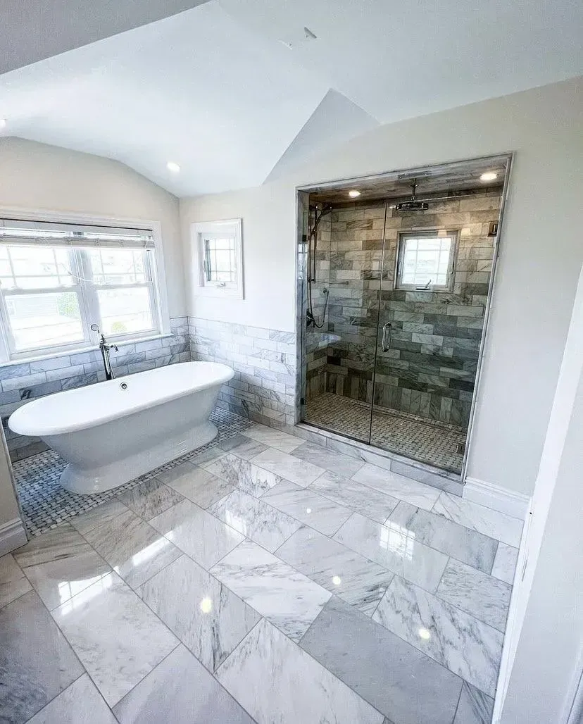 A modern bathroom features a white freestanding tub, a walk-in glass shower with stone tile, and grey marble floor tiles.