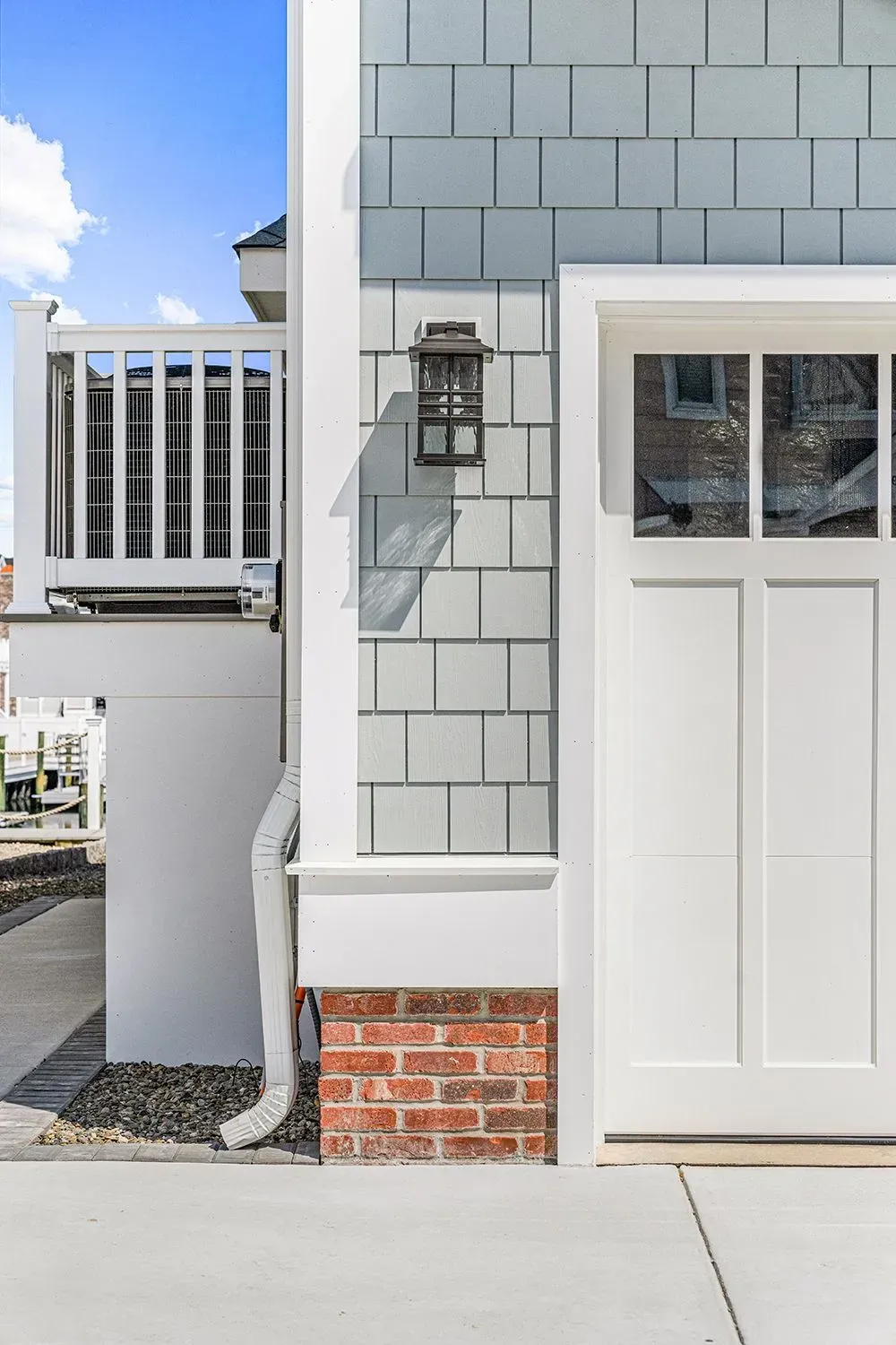 A white garage door next to a gray shingled wall with a lantern, white downspout, and brick base on a bright day.