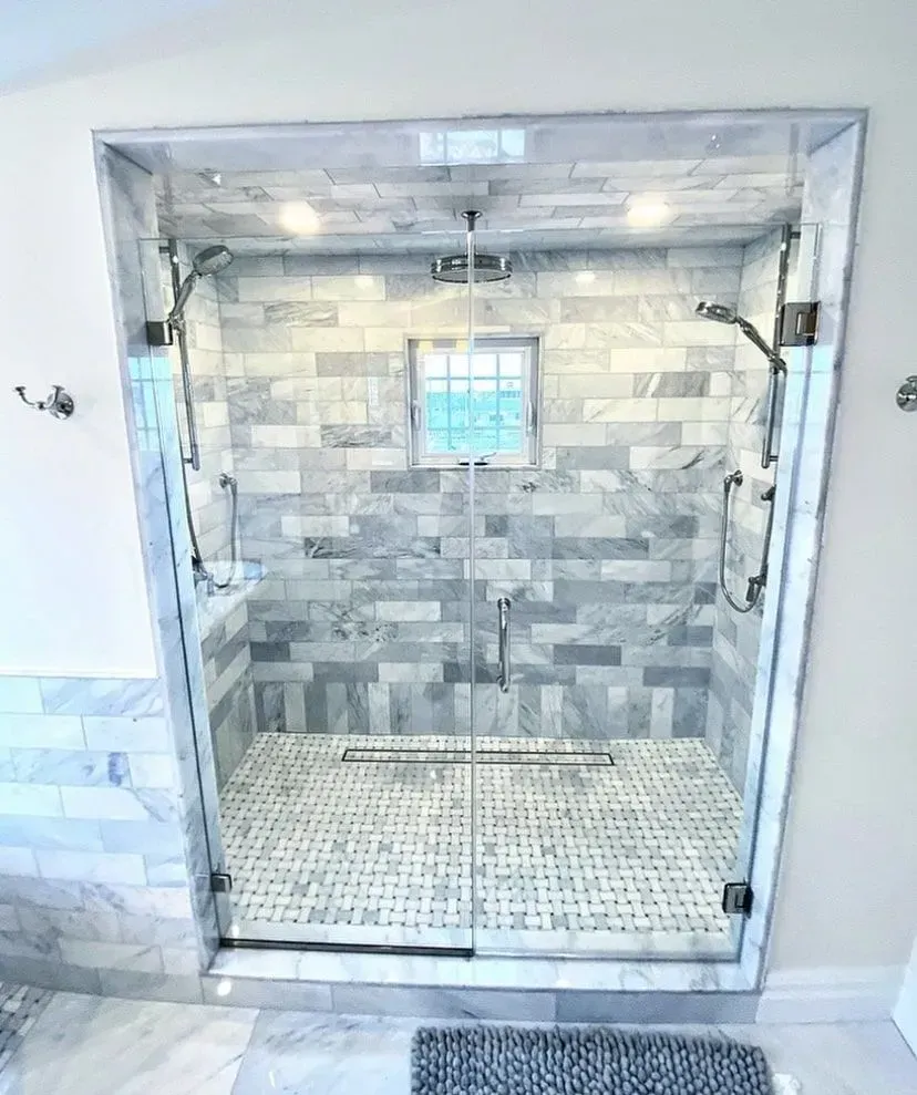 A glass-enclosed walk-in shower with gray marble tile walls, a central window, dual shower heads, and a linear drain.