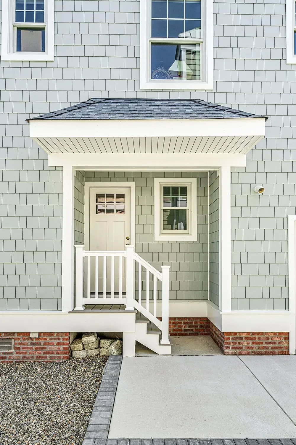 A house entrance with a small covered porch, white stairs, a light-colored door, and gray shingle siding over a brick base.