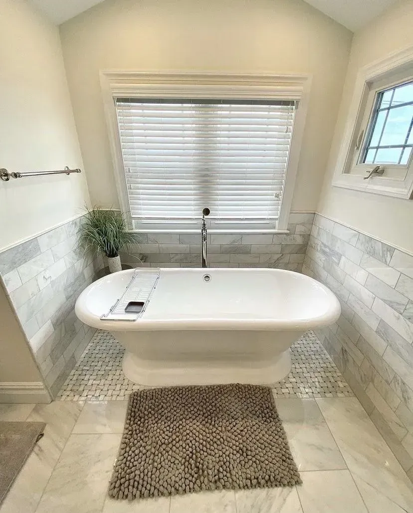 Freestanding white bathtub centered in a marble-tiled bathroom with a window, blinds, and a textured rug on the floor.