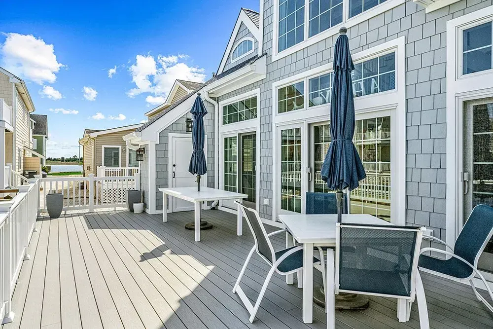 A gray deck with two outdoor dining tables, umbrellas, and chairs, attached to a gray shingled house on a sunny day.