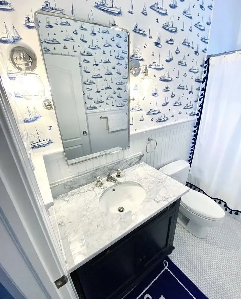 A bathroom vanity with a dark cabinet, white marble countertop, nautical wallpaper, mirror, and white toilet nearby.