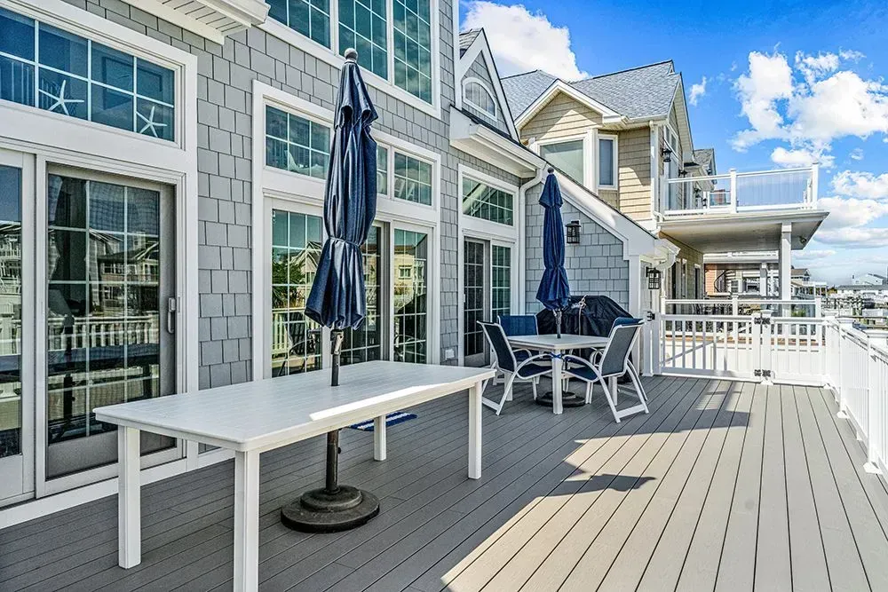 A gray, shingled beach house deck featuring a white table, chairs, and two closed patio umbrellas under a blue sky.