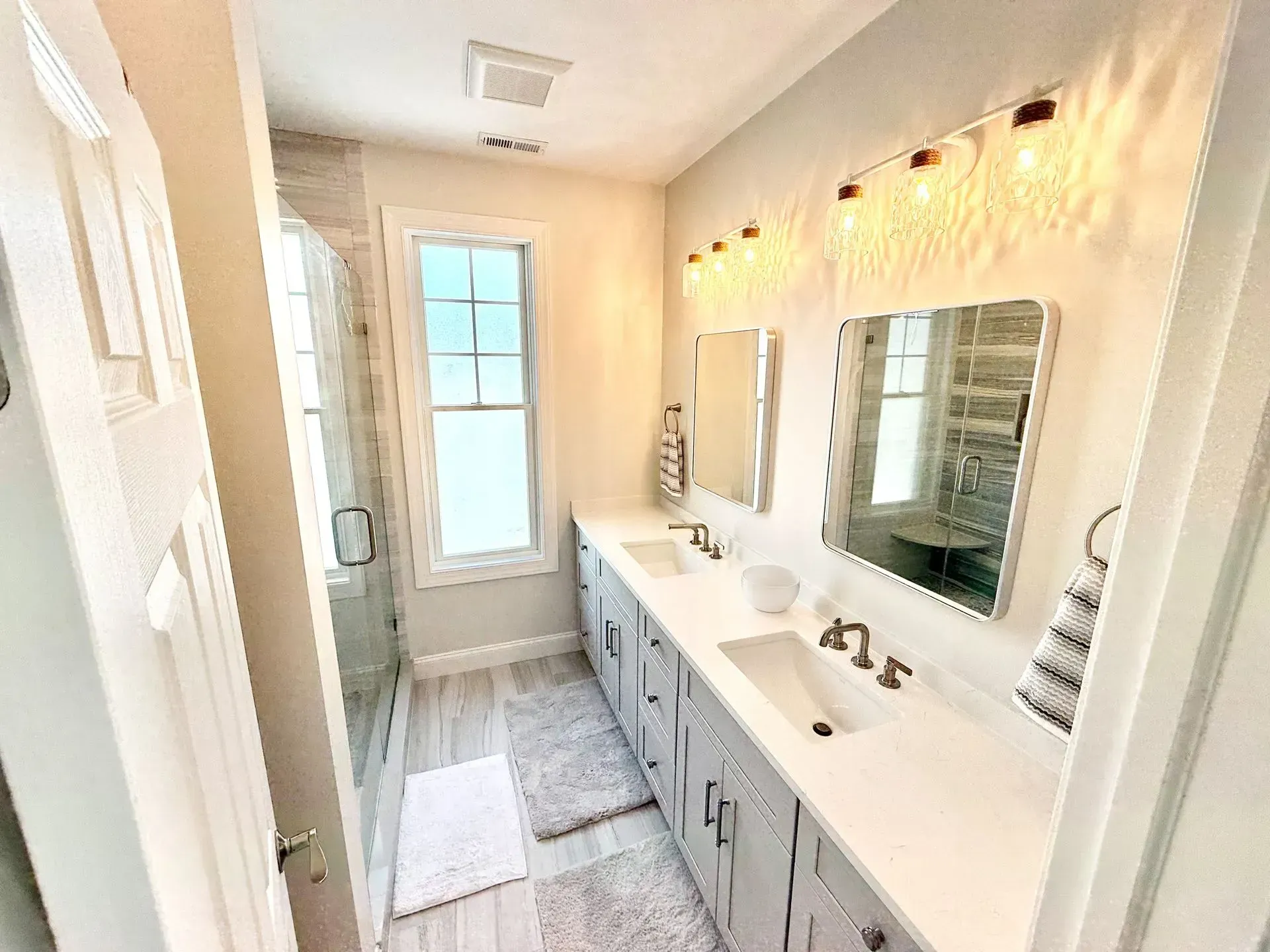 A bright bathroom with a double vanity, gray cabinets, two mirrors, and a glass-enclosed shower near a large window.