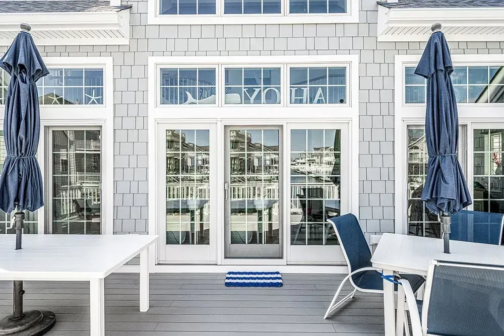 A gray-shingled house exterior with a glass door, white trim, closed blue patio umbrellas, and outdoor chairs on a deck.