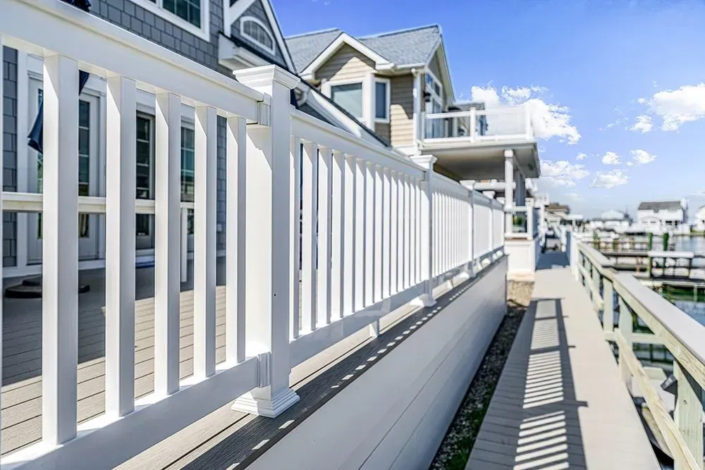 A white deck railing lines a wooden walkway along a canal next to modern houses under a clear blue sky.