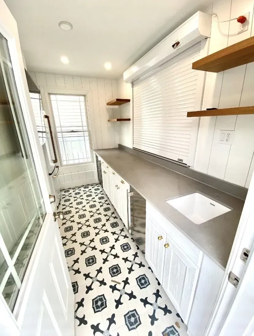 A narrow white kitchen with patterned tile flooring, a gray countertop, a white sink, and wooden floating shelves.