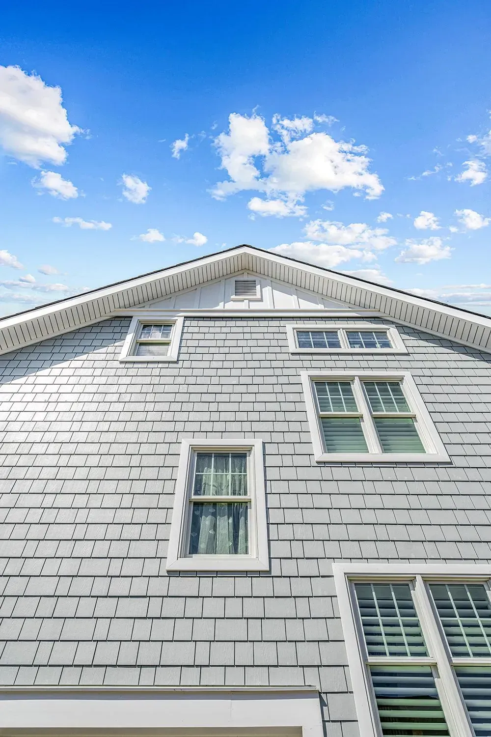 A low-angle view of a grey-shingled house exterior with multiple windows against a blue sky with scattered clouds.