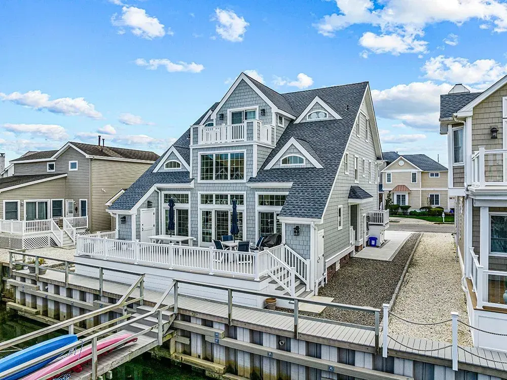 A multi-story gray waterfront house with a white deck, porch, and private dock under a blue sky with scattered clouds.