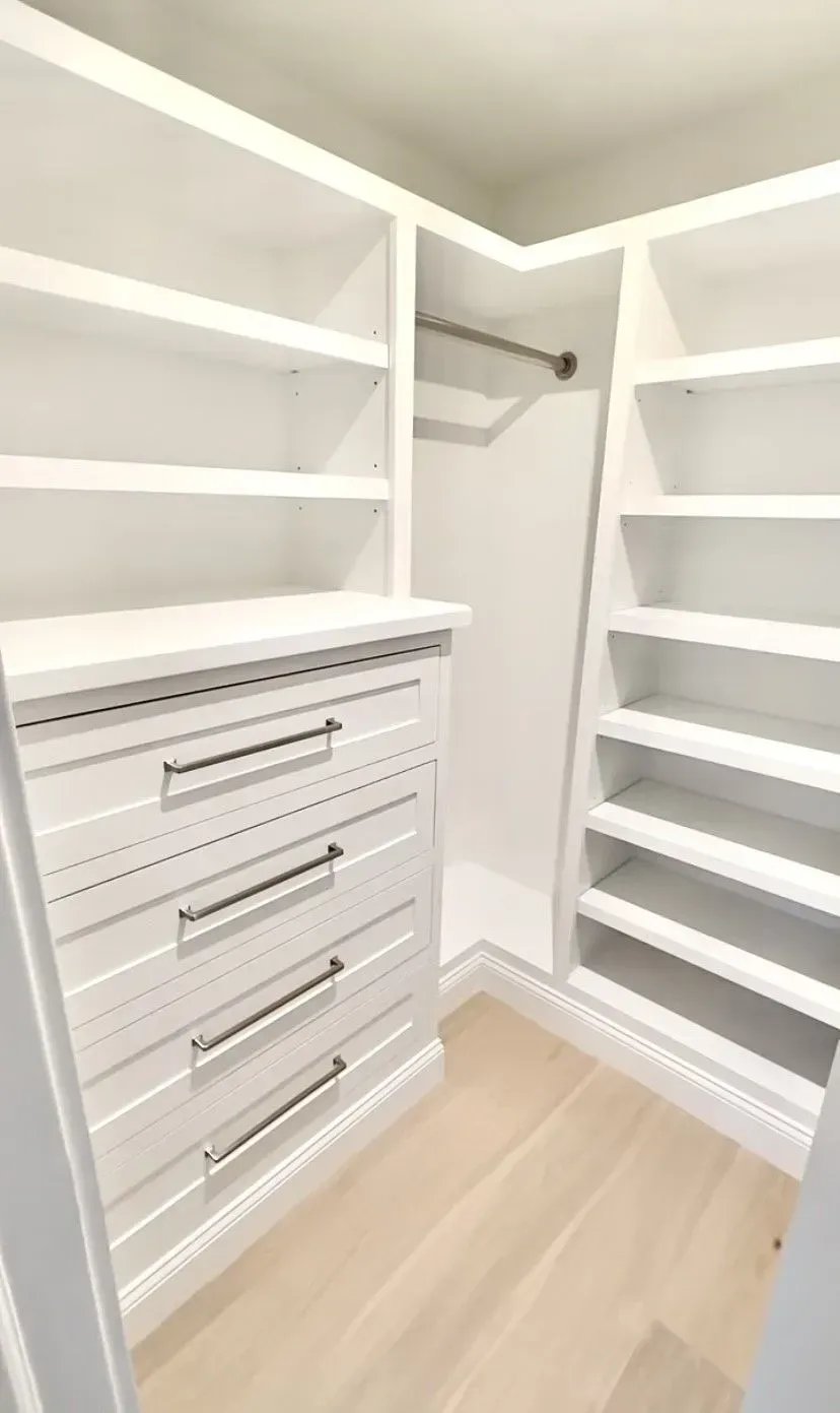 A white, modern walk-in closet featuring built-in shelving, a set of four drawers, and a hanging rod over light wood floors.
