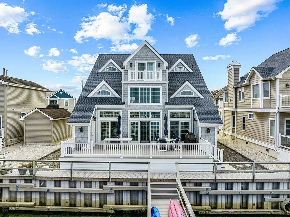 A three-story, grey, waterfront house with a white deck, porch, and a dock extending over the water under a sunny sky.