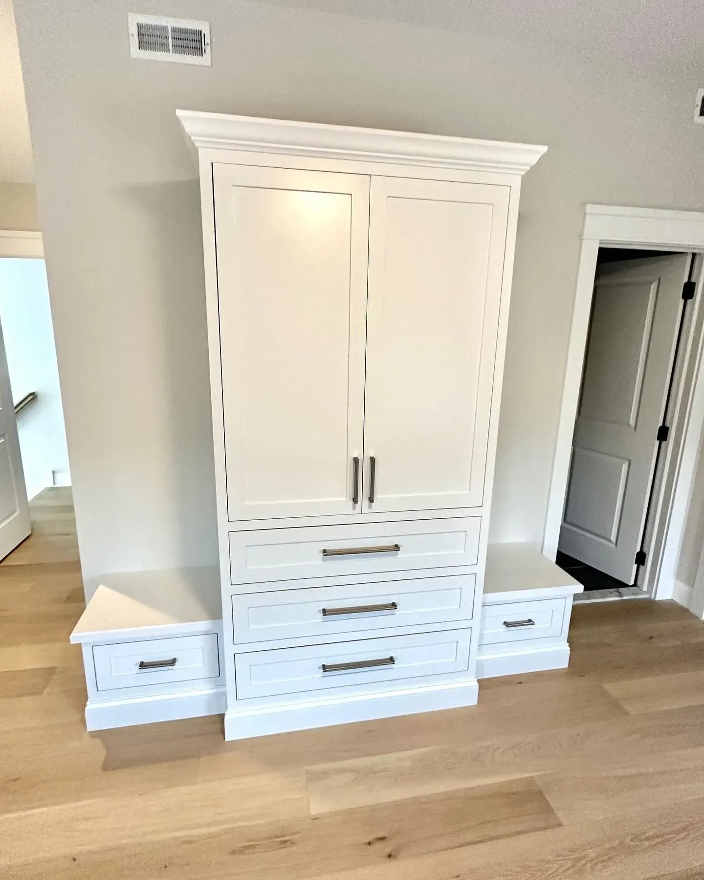 A white cabinet unit featuring two doors above three drawers, flanked by two lower benches with drawers on a wood floor.