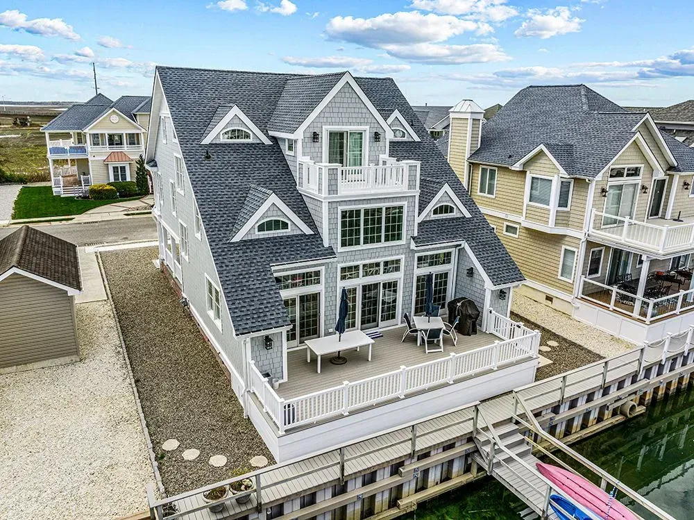 A multi-story gray waterfront house with a deck, gabled roof, and white railings, adjacent to a canal with small boats.