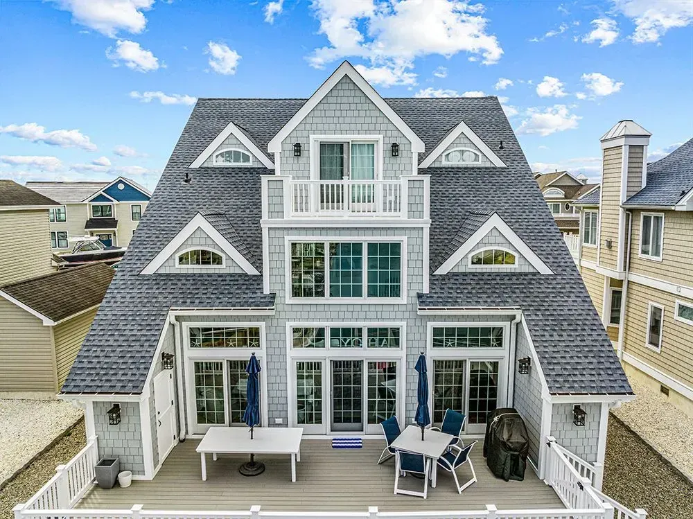 A three-story gray shingled house with a deck, patio furniture, and a balcony under a bright blue sky.