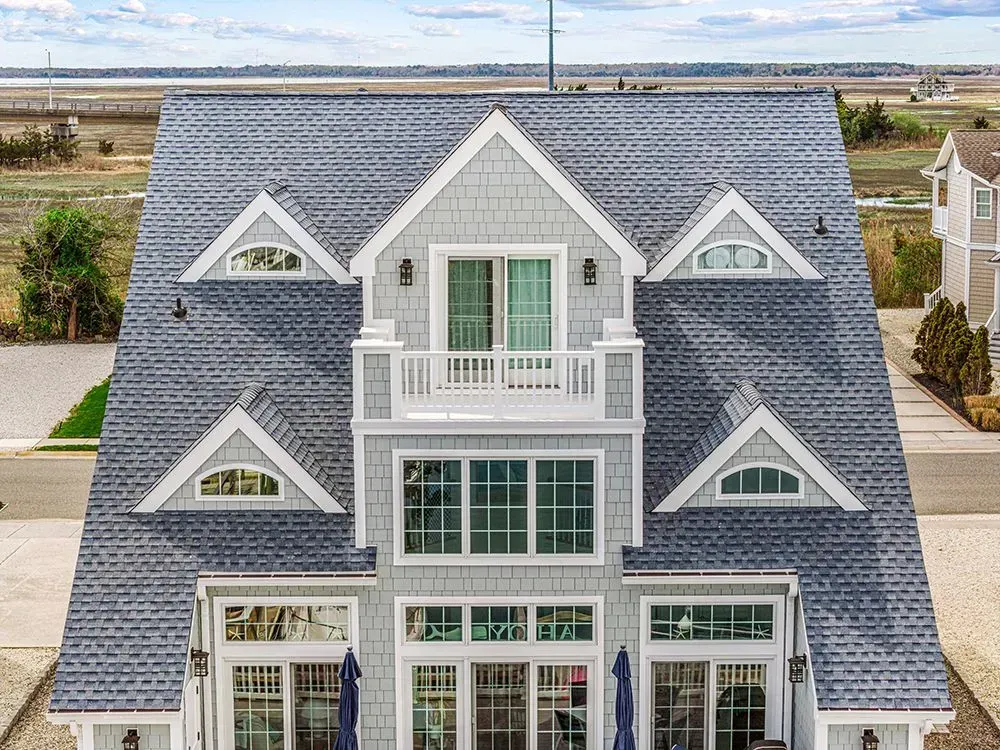A large, three-story, grey shingled house with a steep roof, multiple triangular dormers, and expansive glass doors.