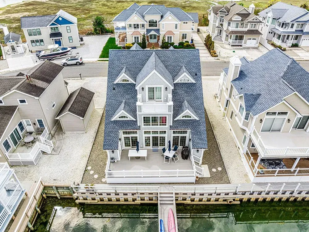 An aerial view of a three-story gray house with a deck overlooking a canal, surrounded by other coastal residential homes.