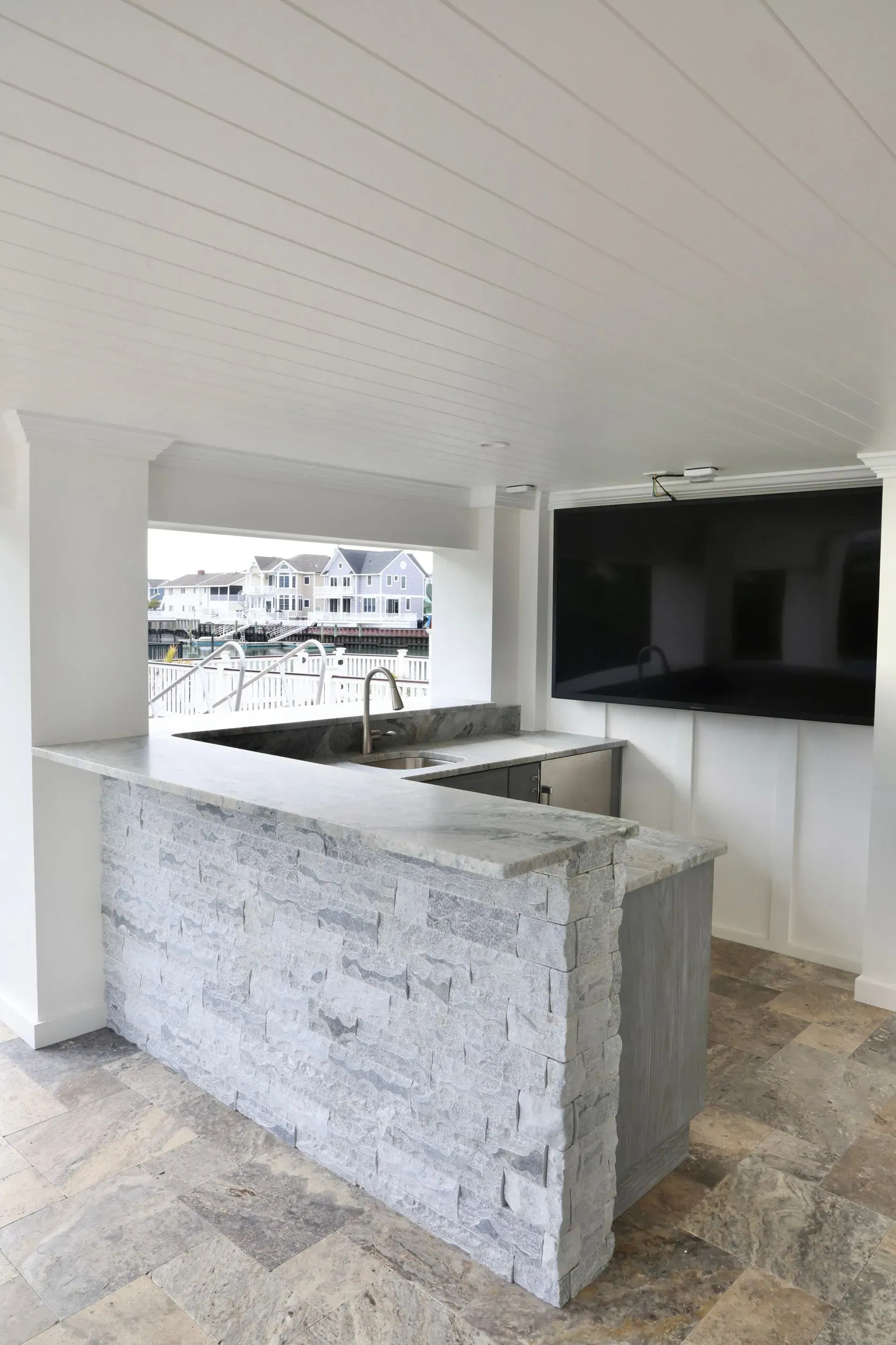 An outdoor bar with a stone base, granite countertop, sink, and wall-mounted television overlooking a marina.