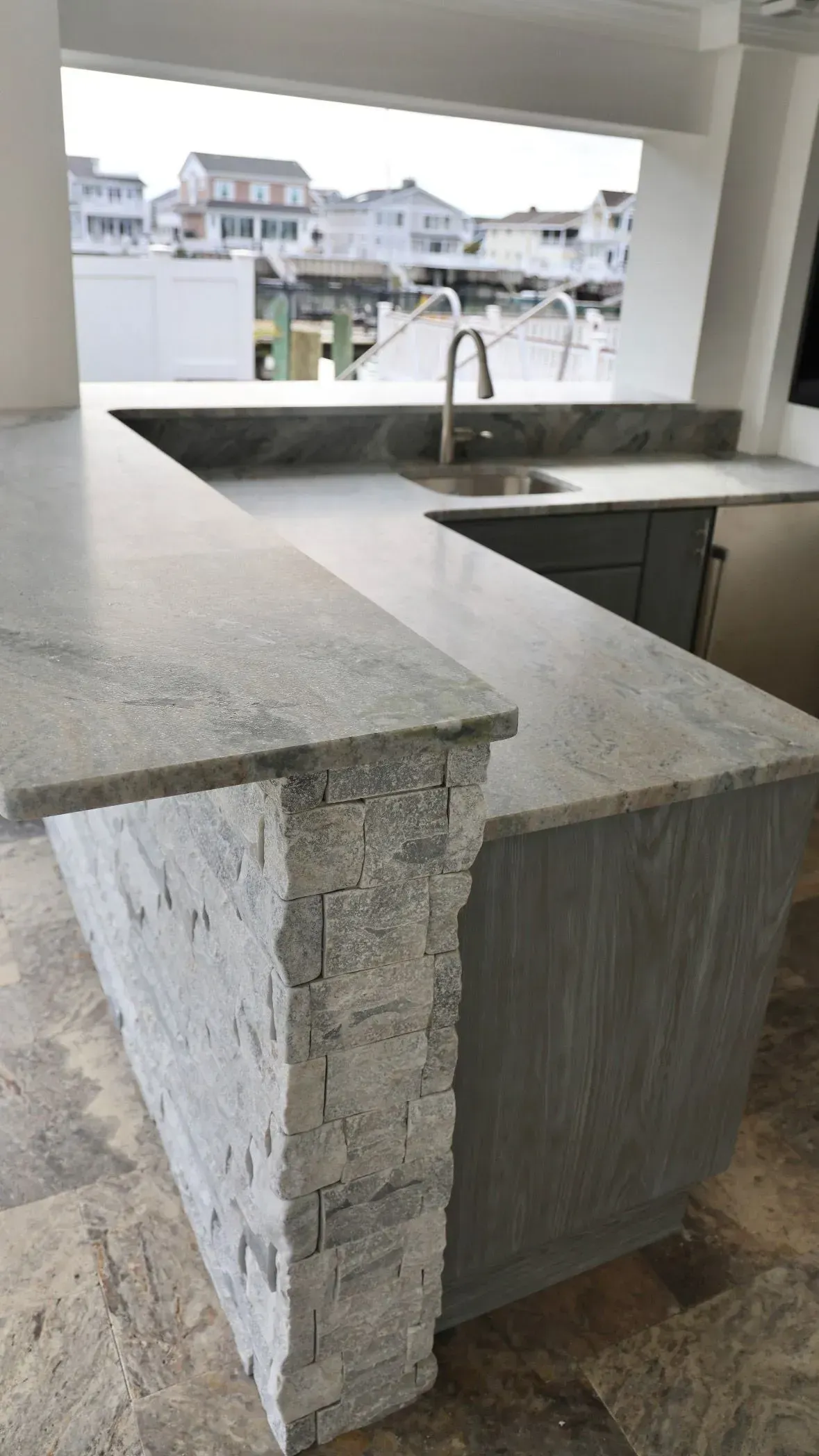An outdoor kitchen bar with a granite countertop, stone facade, and sink, overlooking a marina with houses.