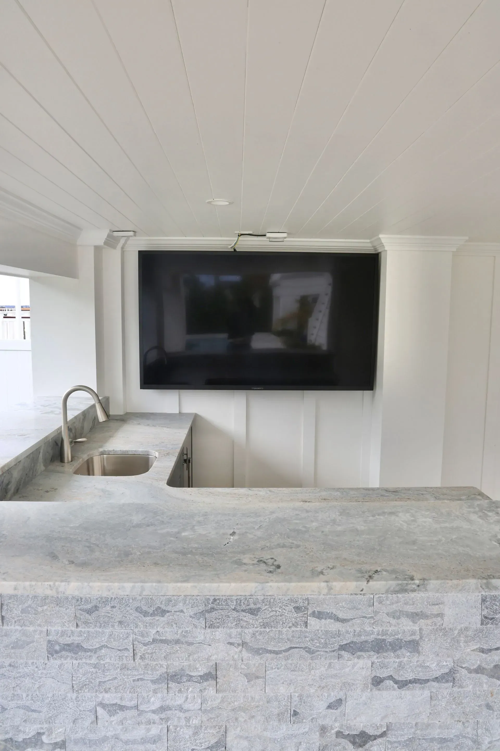 A modern, minimalist outdoor kitchen featuring a grey stone countertop, a small sink, and a large wall-mounted TV.