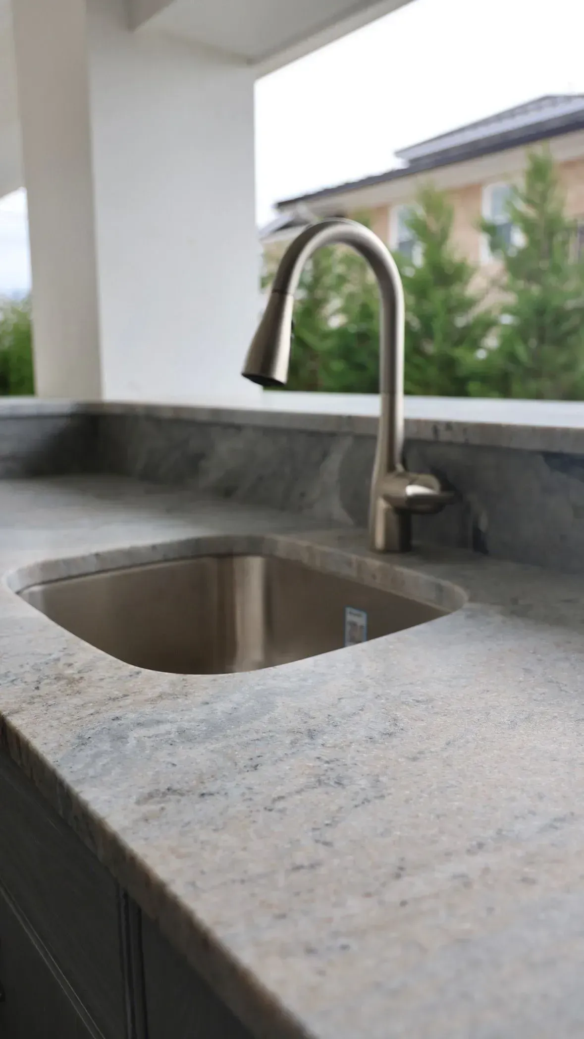 A stainless steel faucet and integrated sink set in a light grey granite countertop, with a blurred outdoor view.