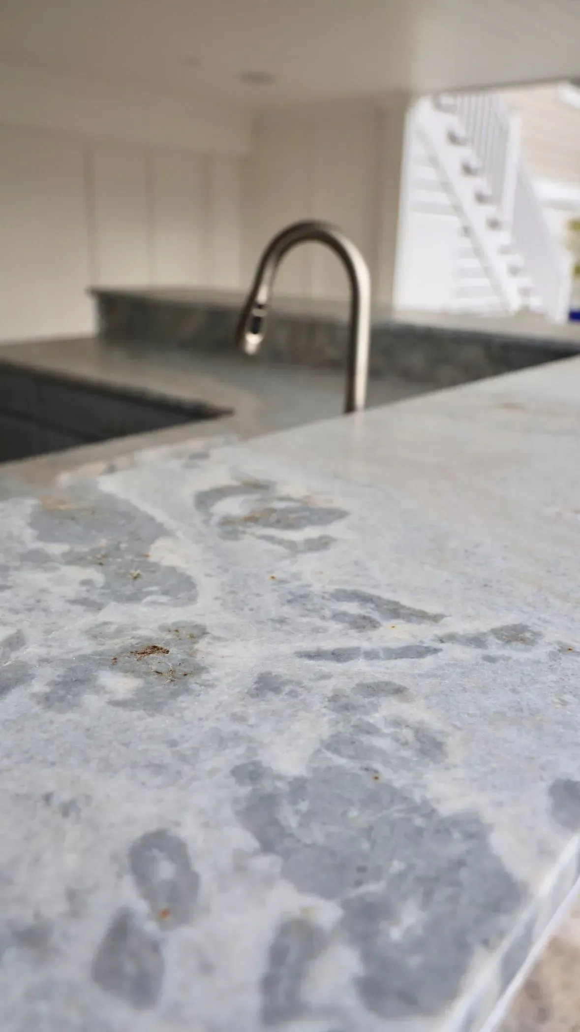 A close-up view of a gray and white marble countertop with a modern, brushed nickel kitchen faucet in the background.
