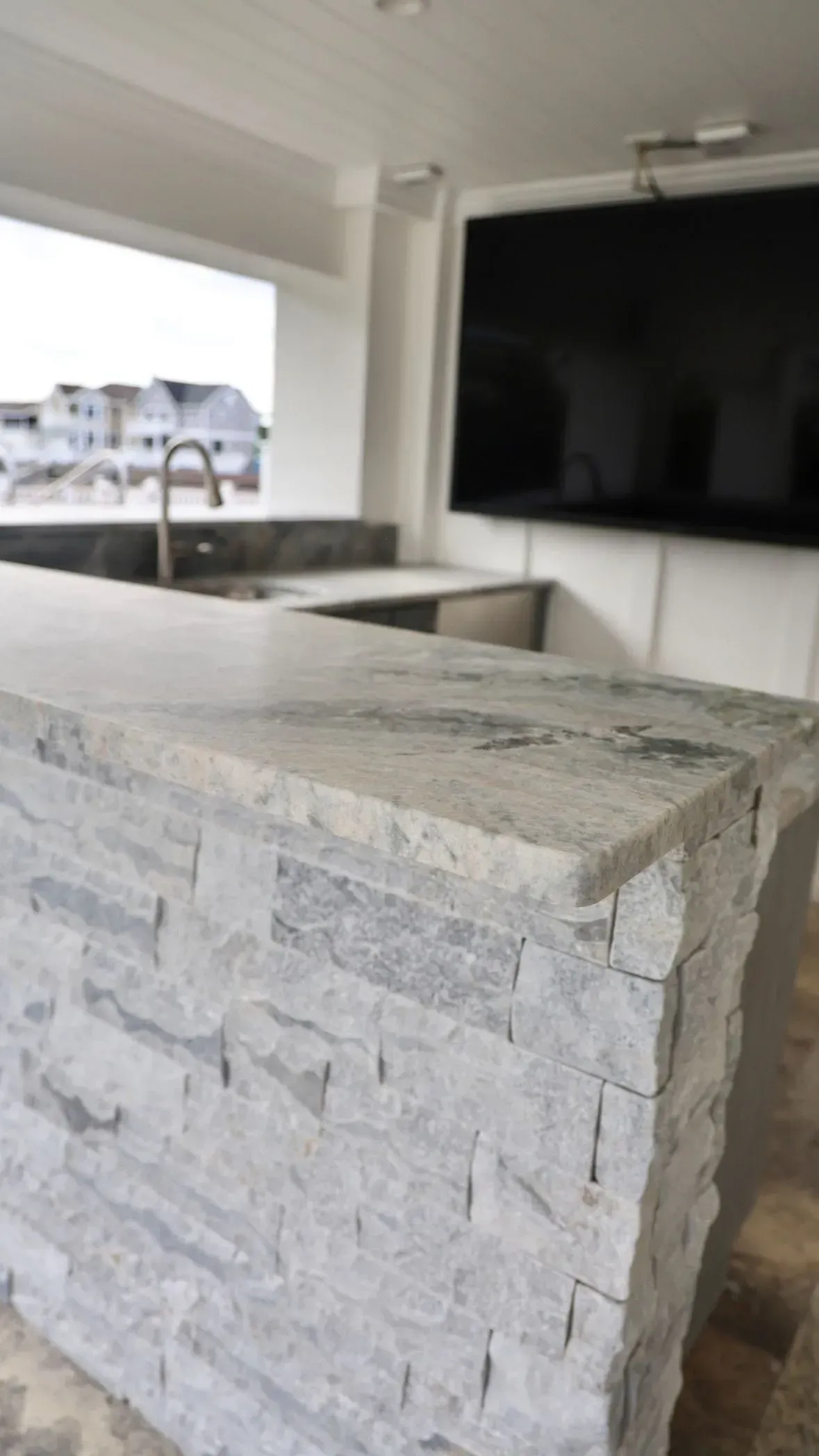 Outdoor bar with a stone facade, granite countertop, sink, and large television mounted on the wall.