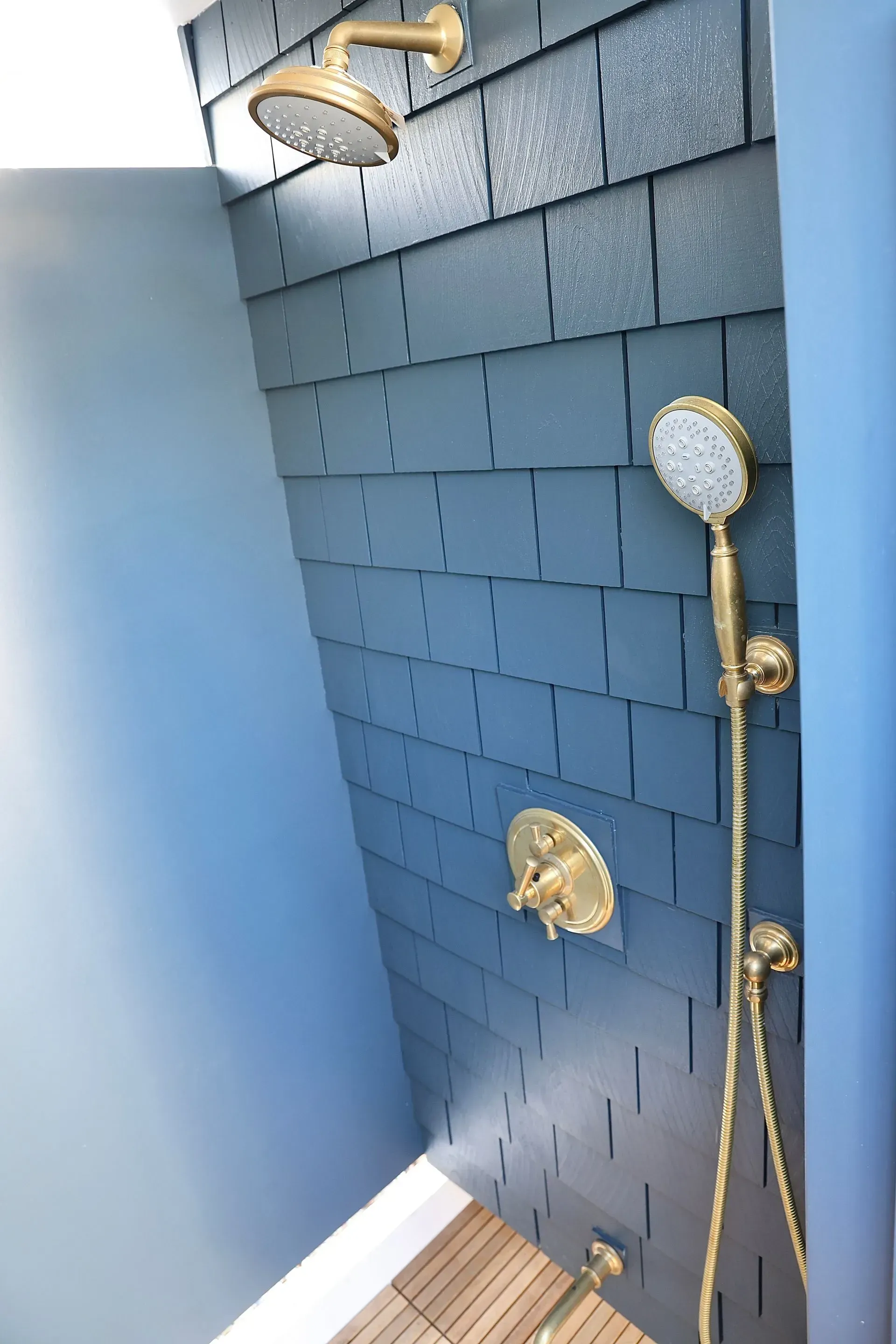 A shower area featuring brass fixtures, a handheld sprayer, and a rain showerhead mounted on a dark blue shingled wall.