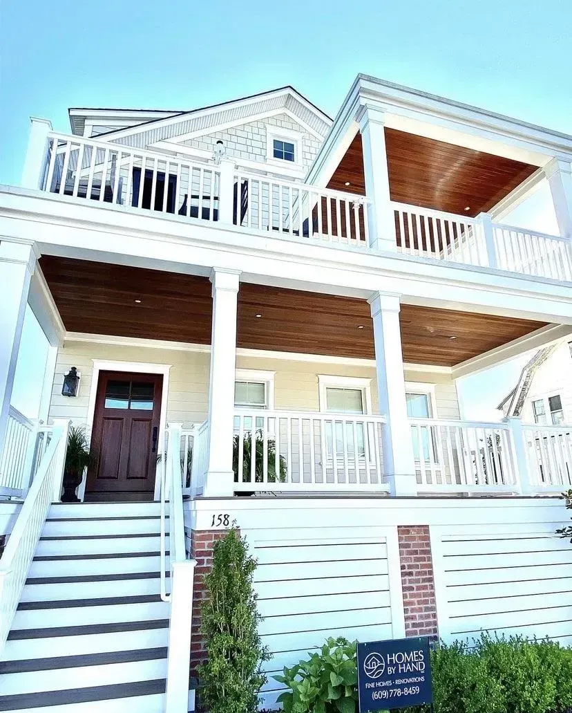 A light-colored two-story beach house with a prominent white porch, dark wood ceilings, and a staircase entry.